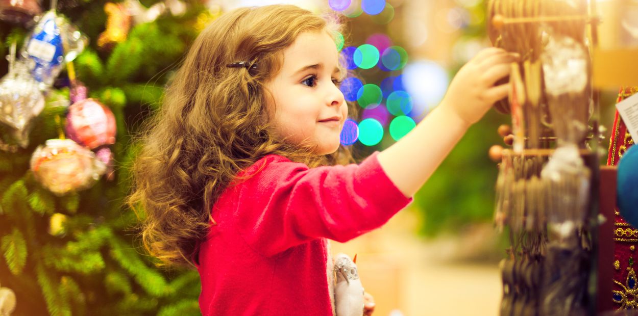 A young brown haired girl is wearing a red shirt. She's standing in front of a decorated Christmas tree and her arm is outstretched to choose an ornament from a store display